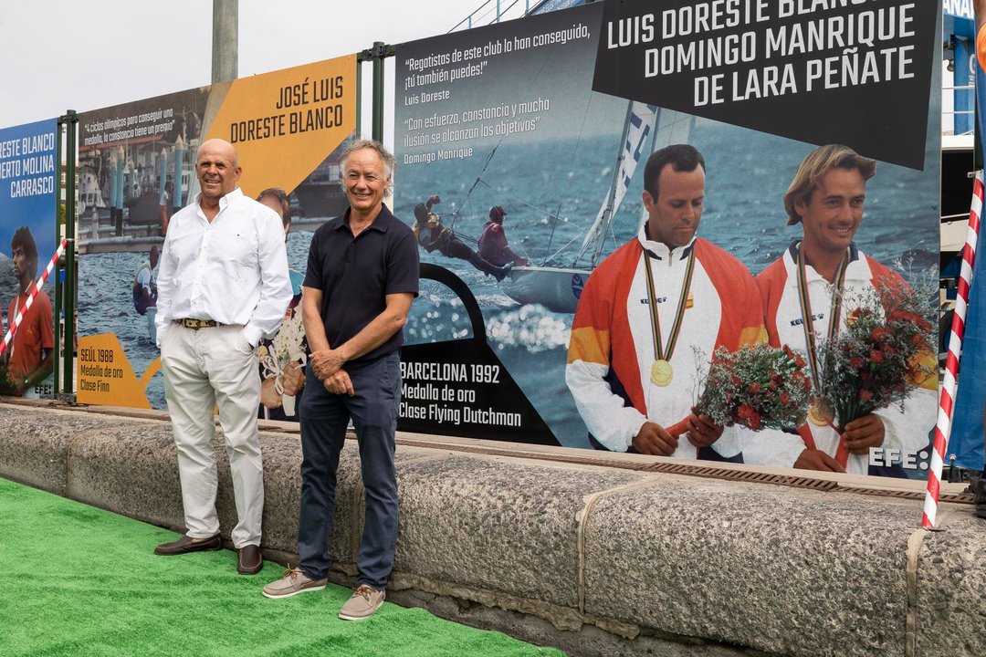 Los regatistas Luis Doreste (d) y Domingo Manrique (i), medallas de oro de vela en los Juegos de Barcelona de 1992 en la clase flying dutchman, posan ante una foto suya de EFE en el mural que el Real Club Náutico de Gran Canaria ha instalado en la rampa donde embarcan sus tripulaciones para inspirar sus su jóvenes promesas con los logros de sus olímpicos. EFE/Quique Curbelo